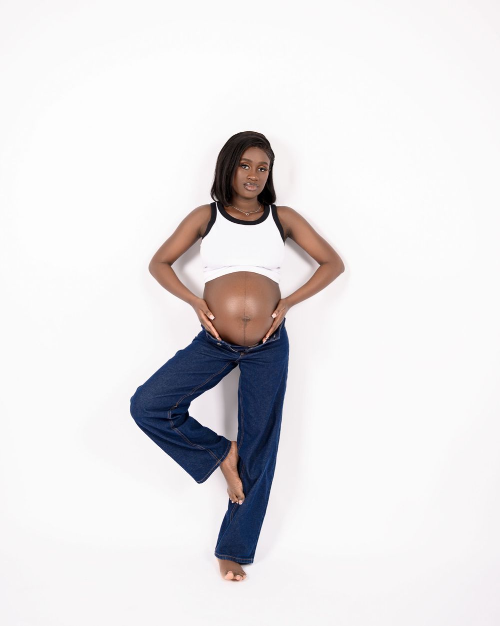 Pregnant person in a white tank top and navy blue pants stands in a dance-like pose against a white backdrop.