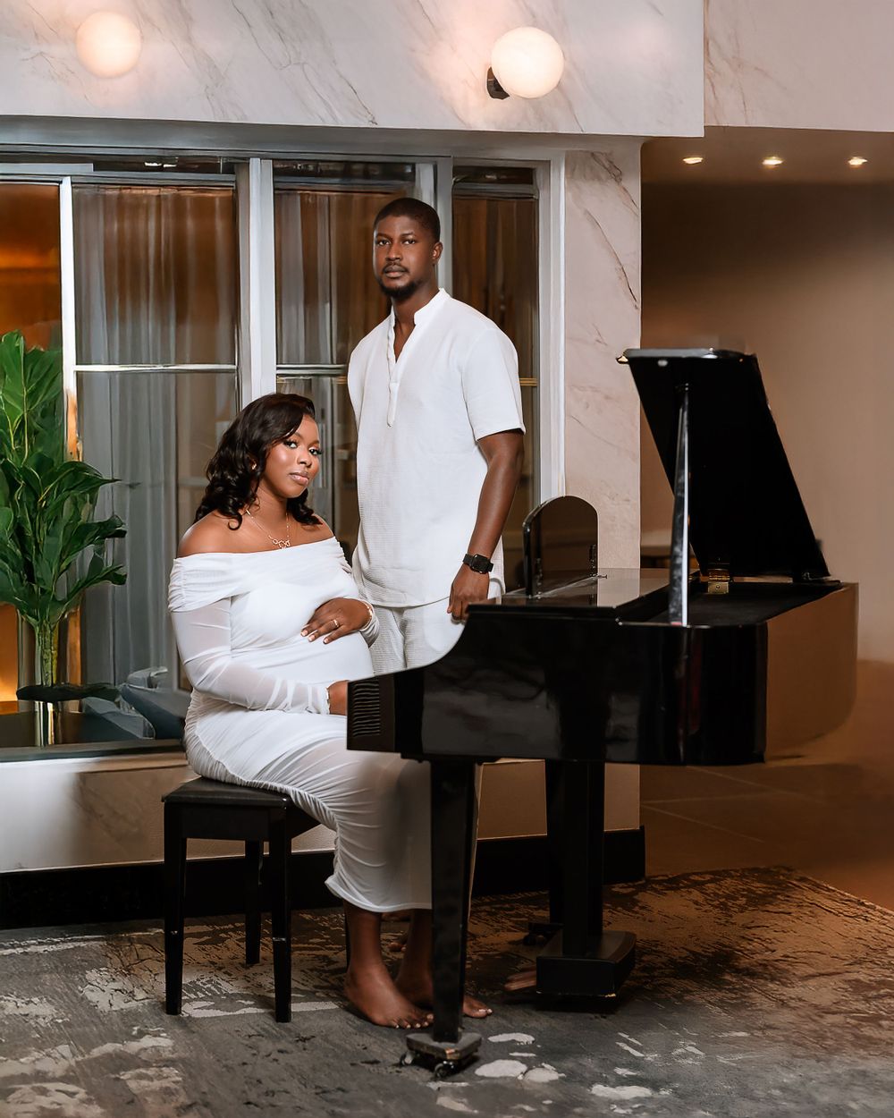 A couple in matching white outfits pose elegantly beside a grand piano in a modern interior setting with warm lighting.