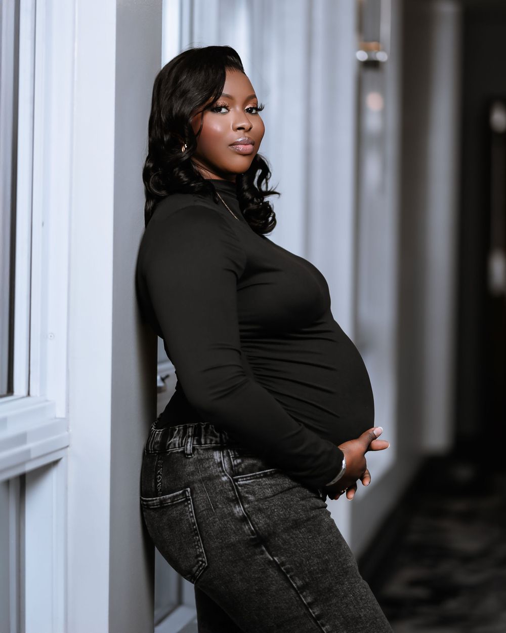 Woman in black turtleneck and jeans poses for a maternity photo against a light-colored wall.