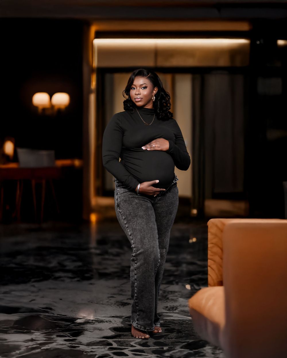 Stylish maternity photo of a person in a black turtleneck and jeans standing in a dimly lit elegant interior space.