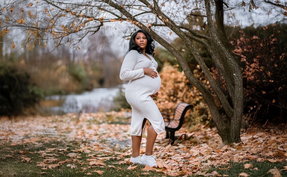 A figure in a cozy white sweater dress and sneakers poses gracefully among autumn leaves in a natural outdoor setting.