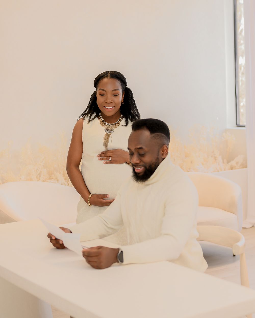 A couple wearing matching cream-colored outfits smiles together in a bright minimal interior setting.