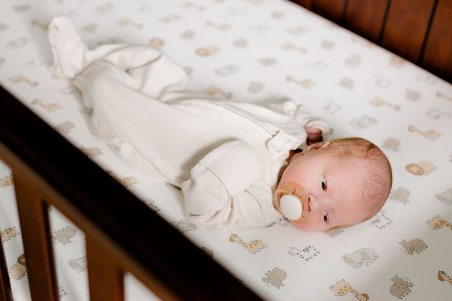 Newborn baby in white outfit with pacifier lying in a dark wood crib with a neutral animal print sheet.
