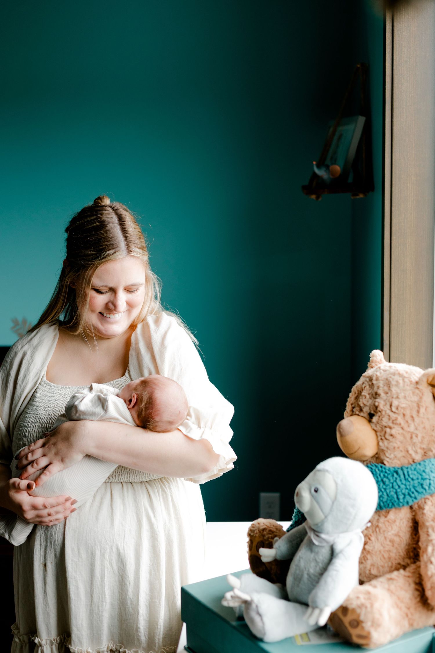 Smiling new mother in cream dress holding newborn baby beside large teddy bears in teal nursery.