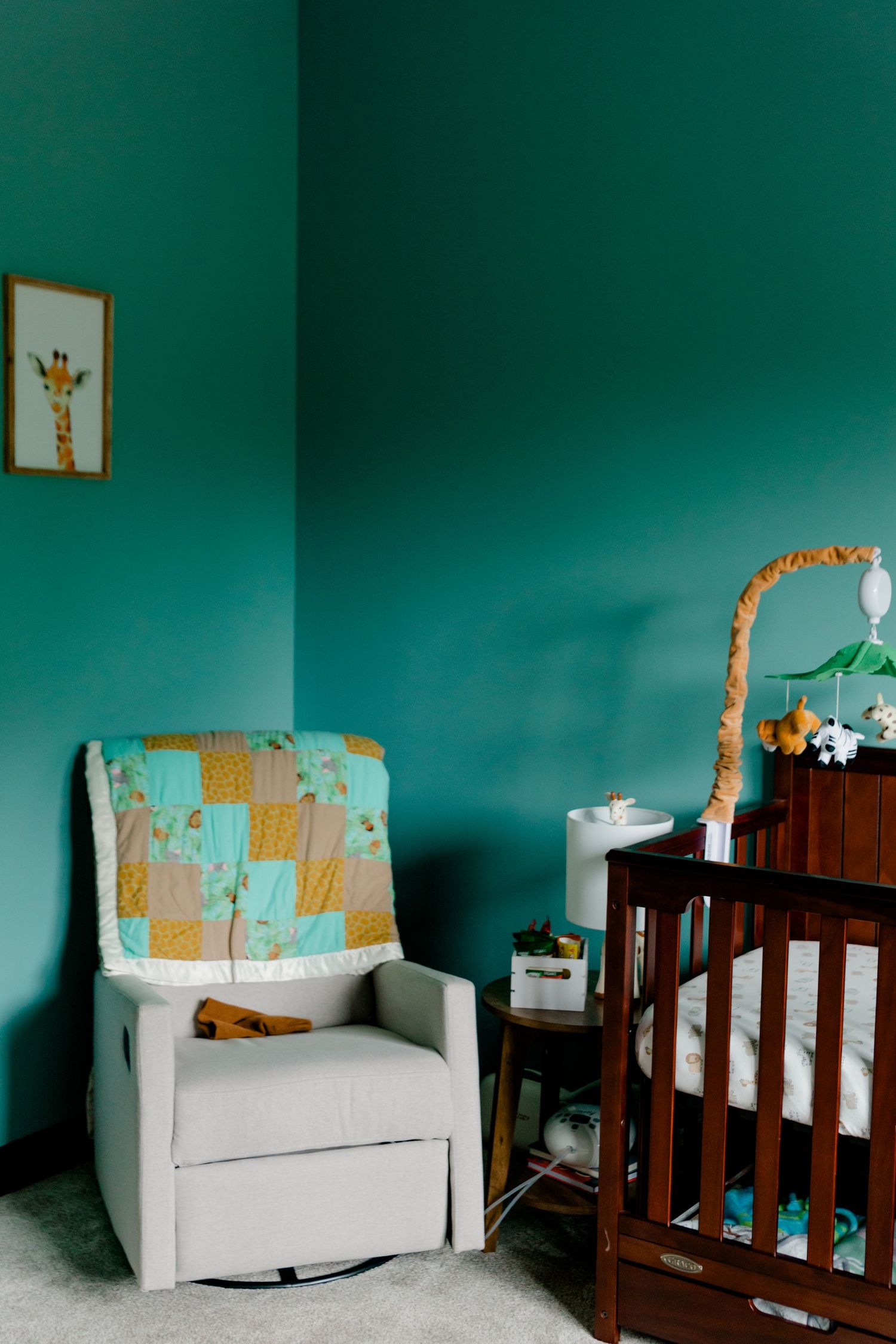 Teal nursery corner with glider chair, giraffe quilt, wooden crib, and safari mobile.