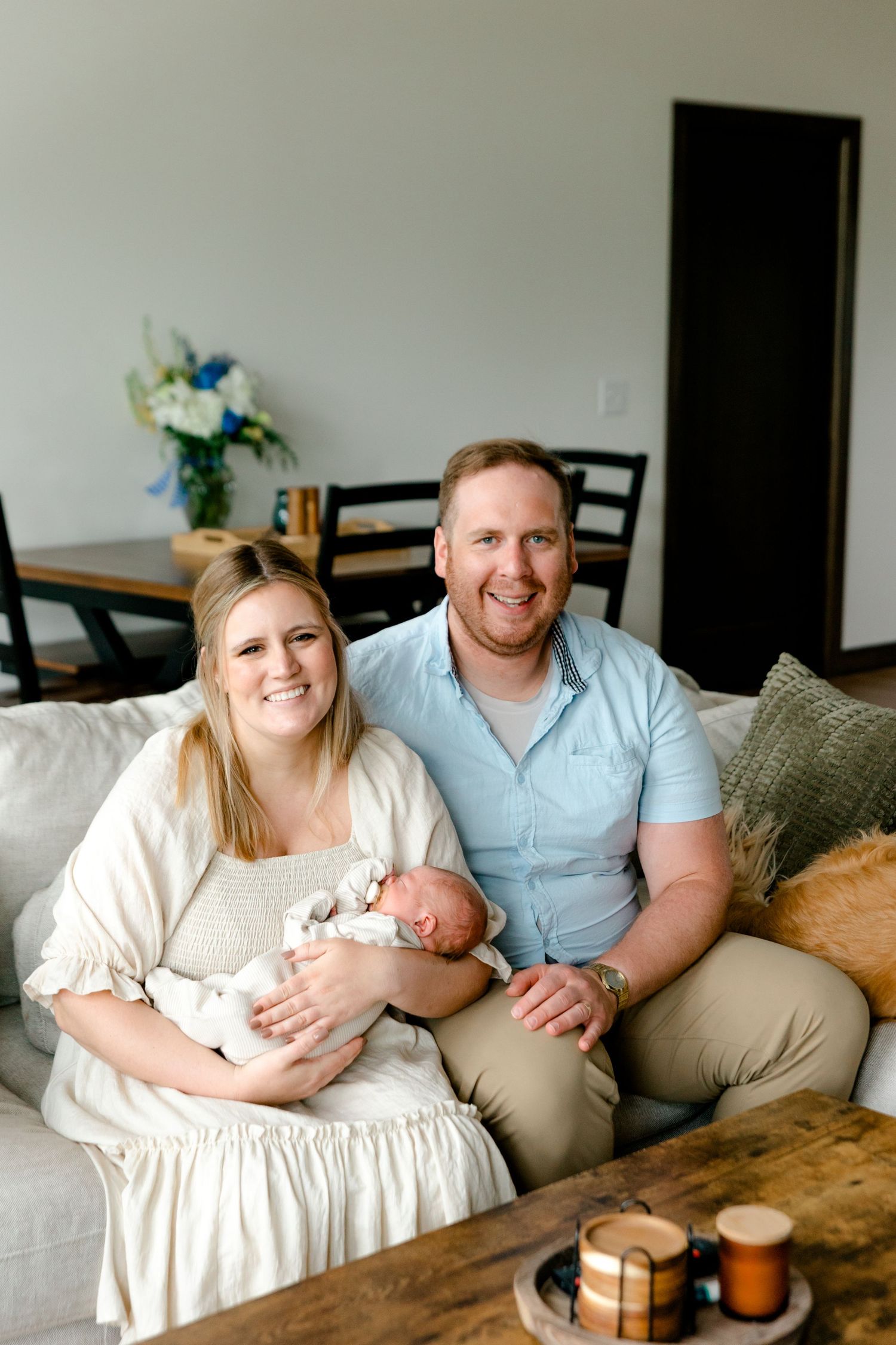 Happy new parents smiling on couch holding sleeping newborn baby in bright modern living room.