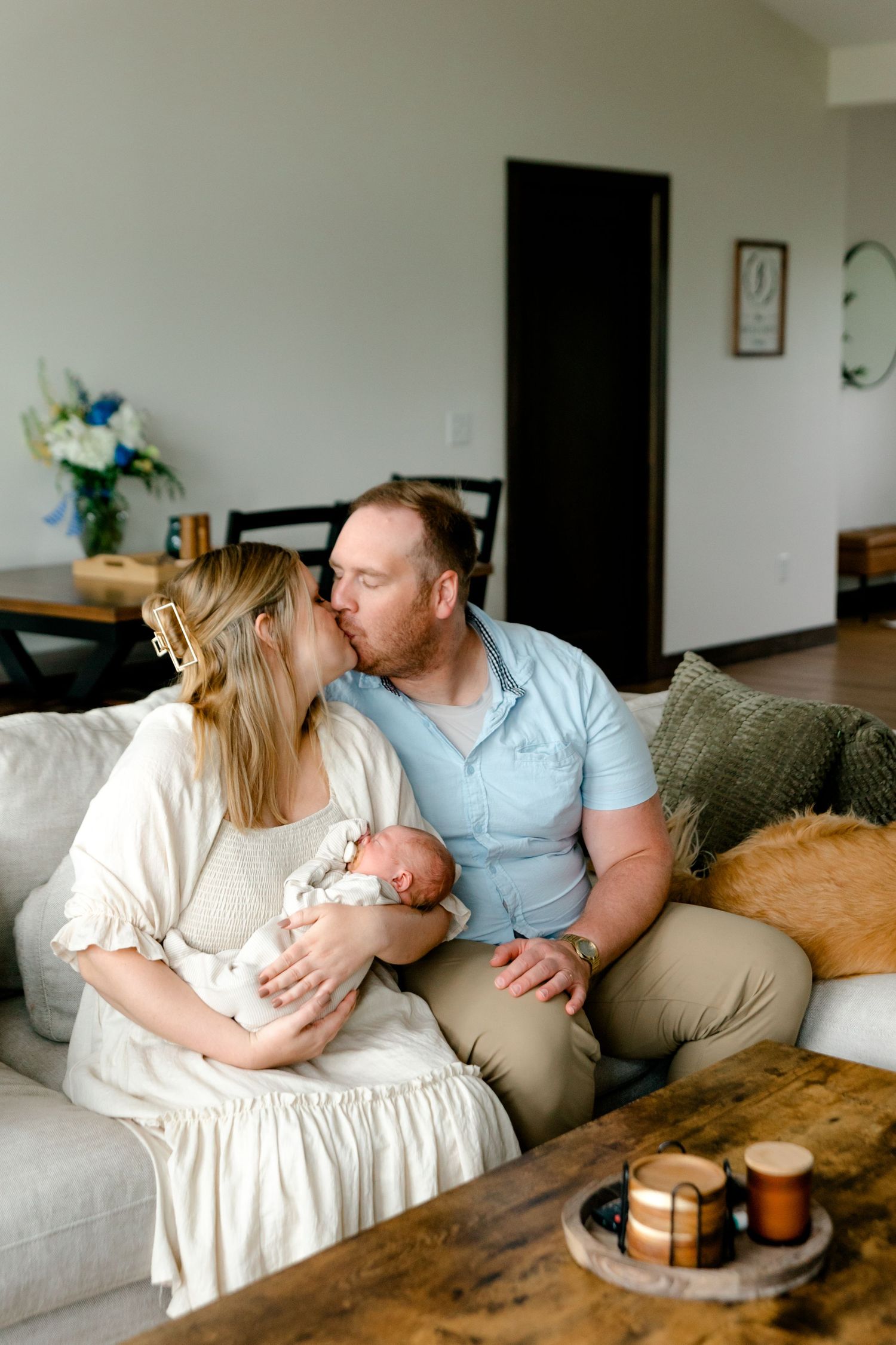 A couple shares a kiss on a couch while holding their newborn baby in a cozy living room.