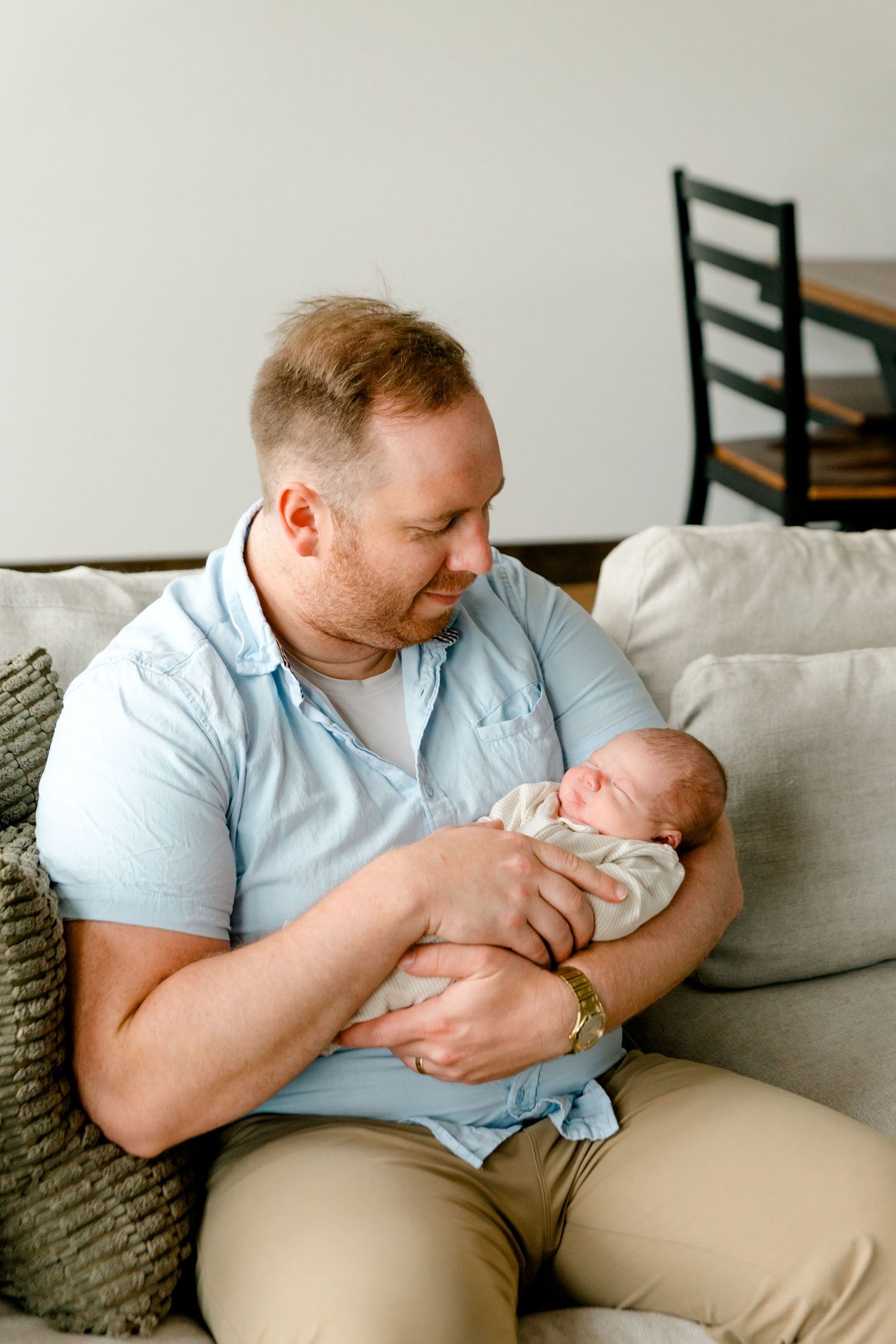 A father in a blue shirt tenderly holds his newborn baby on a couch.