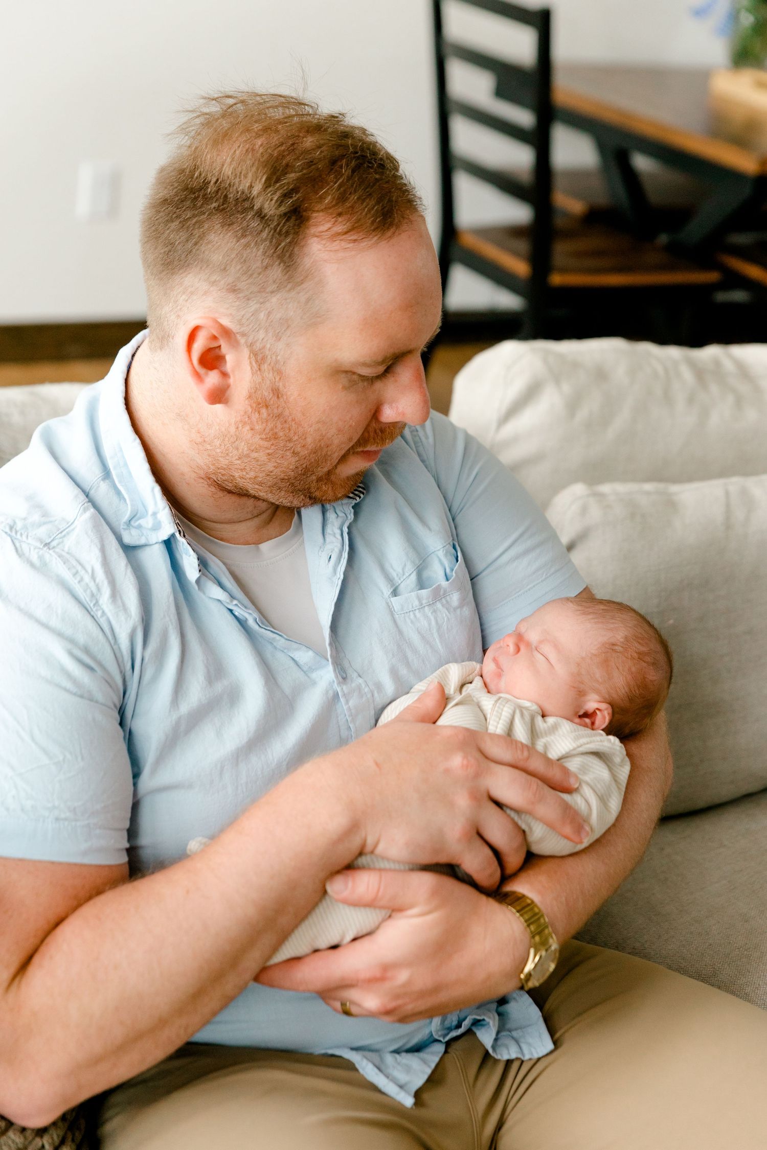 Proud father sitting on a sofa gazing lovingly at his sleeping newborn baby.