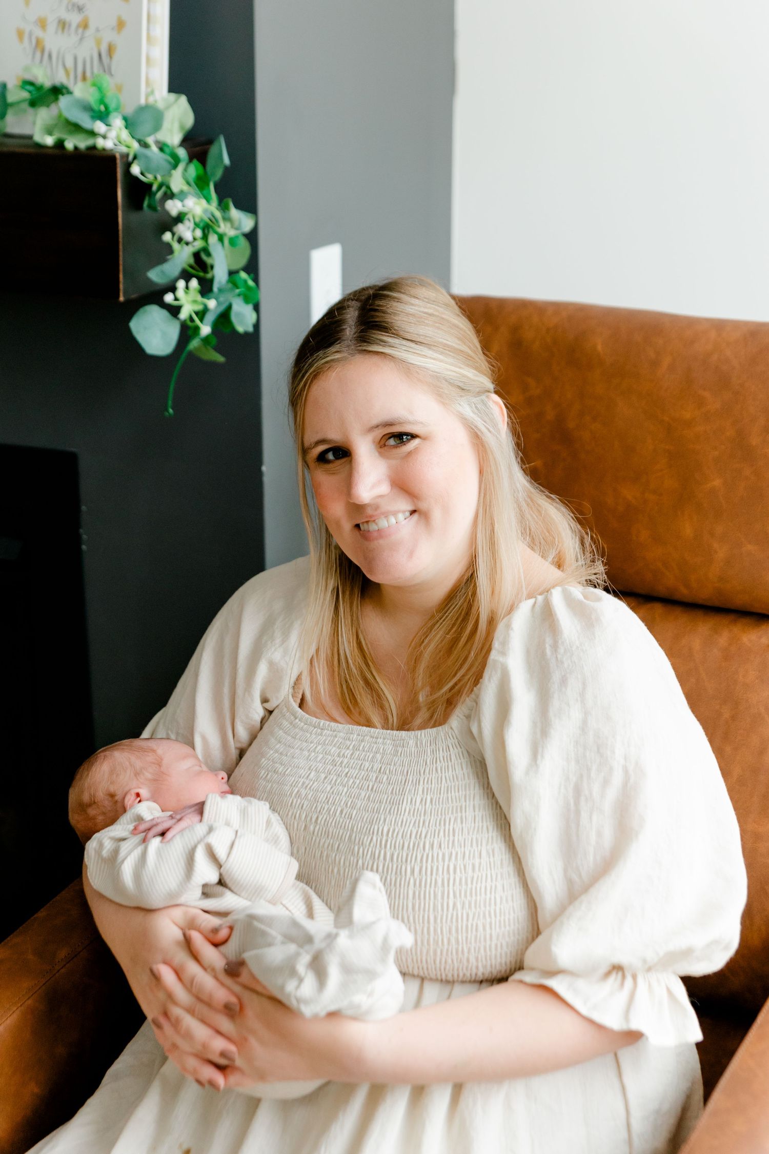 Smiling blonde mother in cream dress holding newborn baby in cozy home setting.