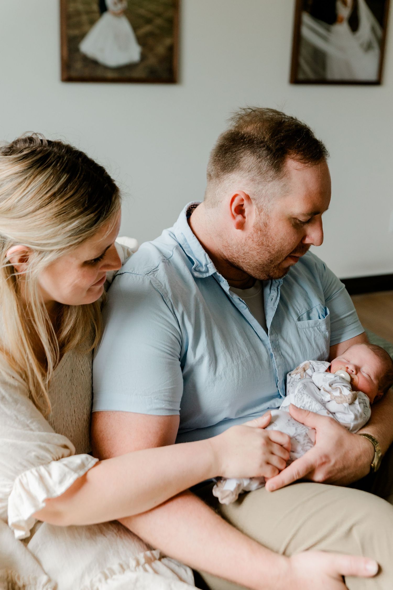 New parents lovingly gazing at their newborn baby with wedding photos on wall behind.