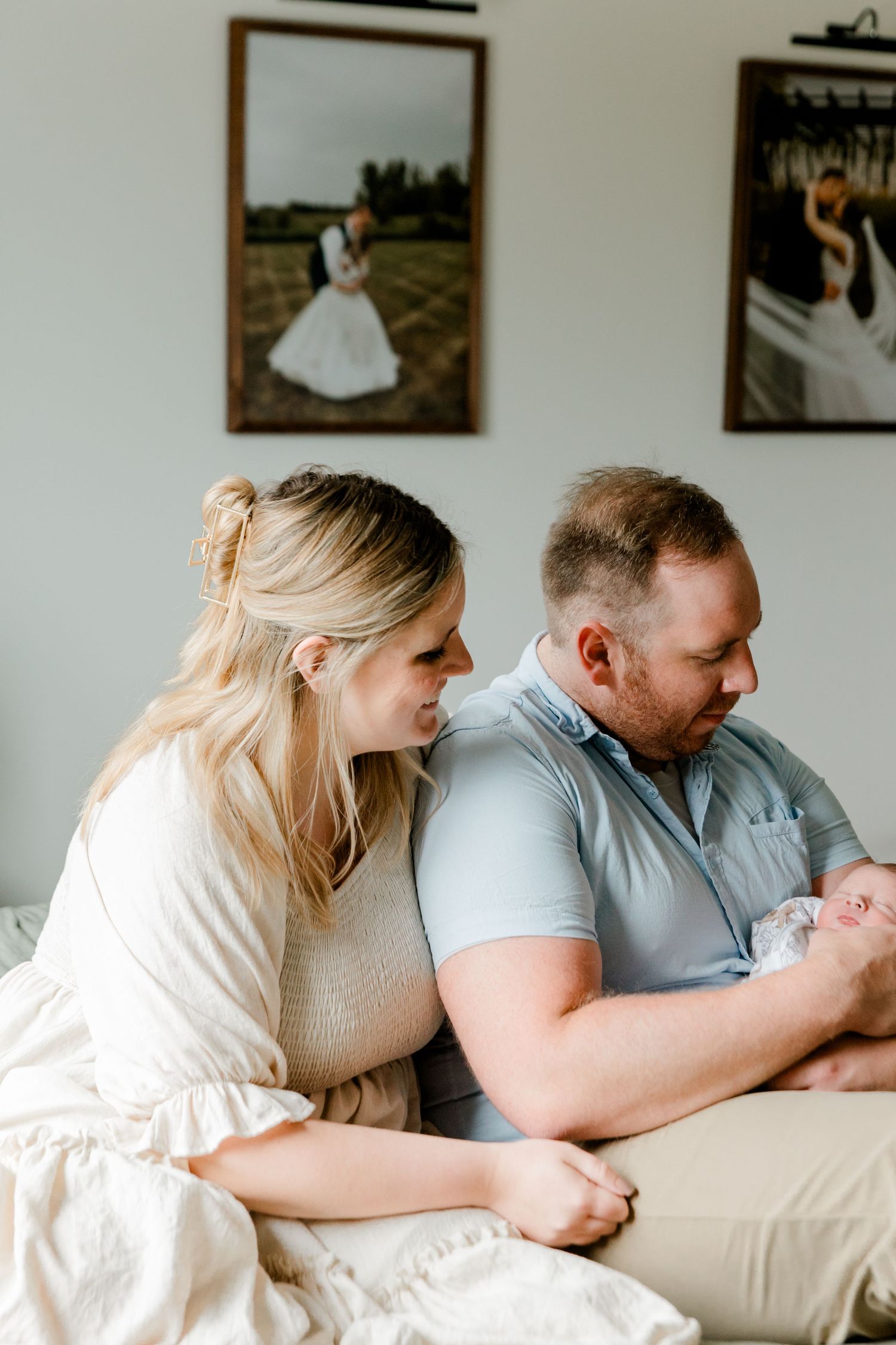 New parents leaning in together to admire their newborn baby with wedding photos behind.