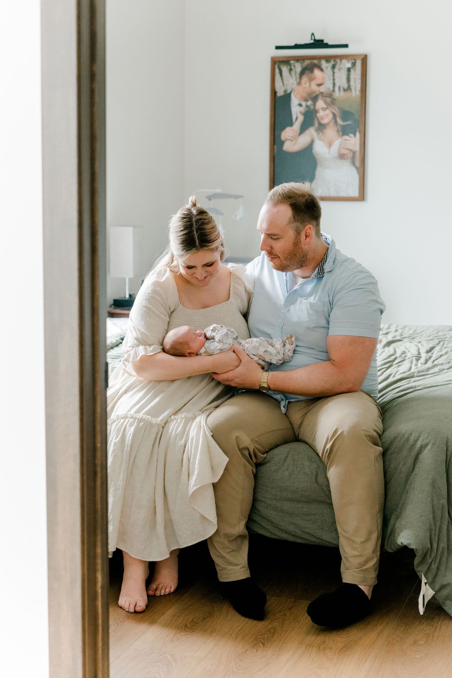 New parents sit on a bed holding their newborn baby, with a wedding photo displayed on the wall behind them.