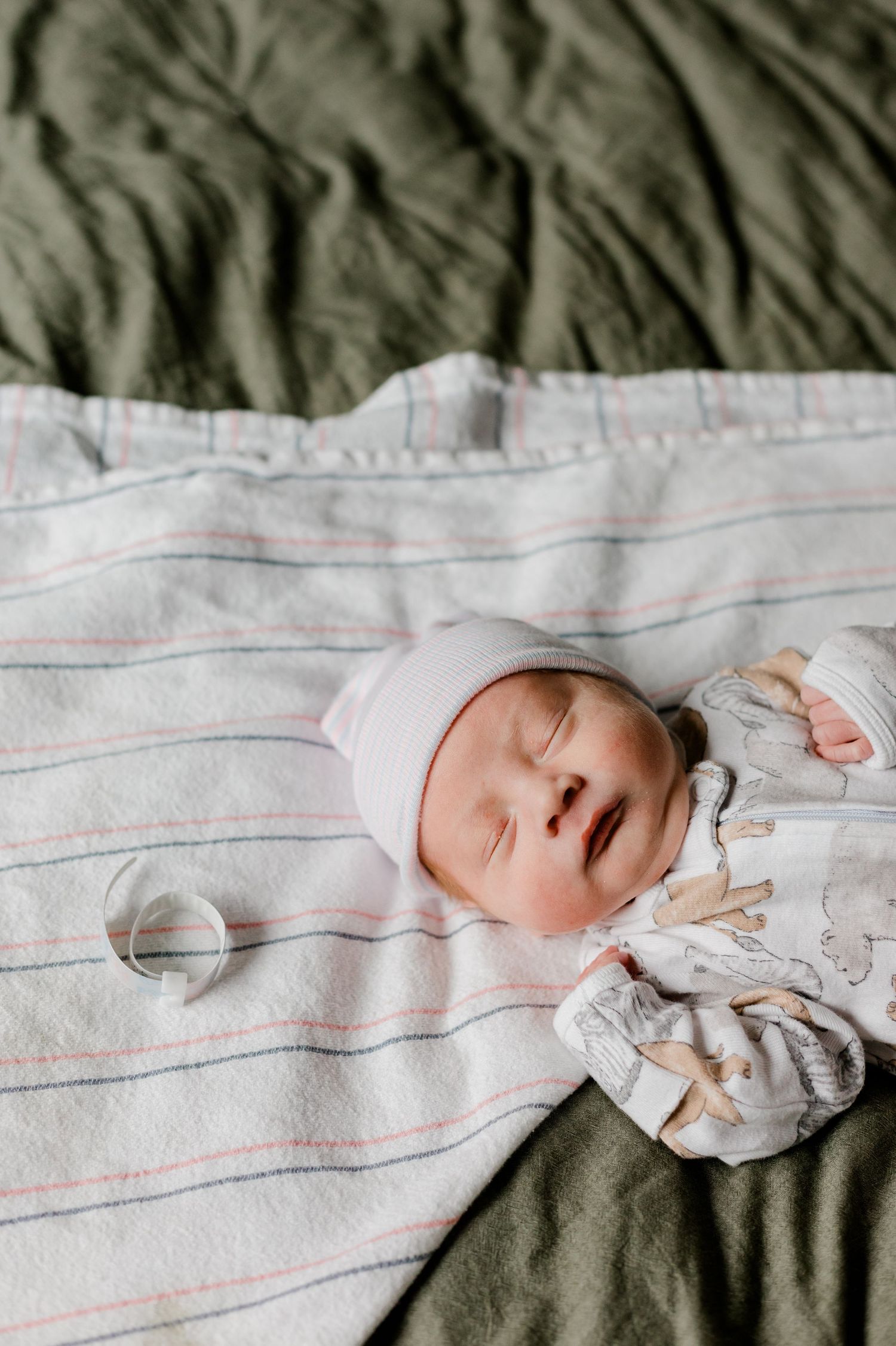 A sleeping newborn baby in a white hat and onesie rests on a striped blanket with a ring nearby.