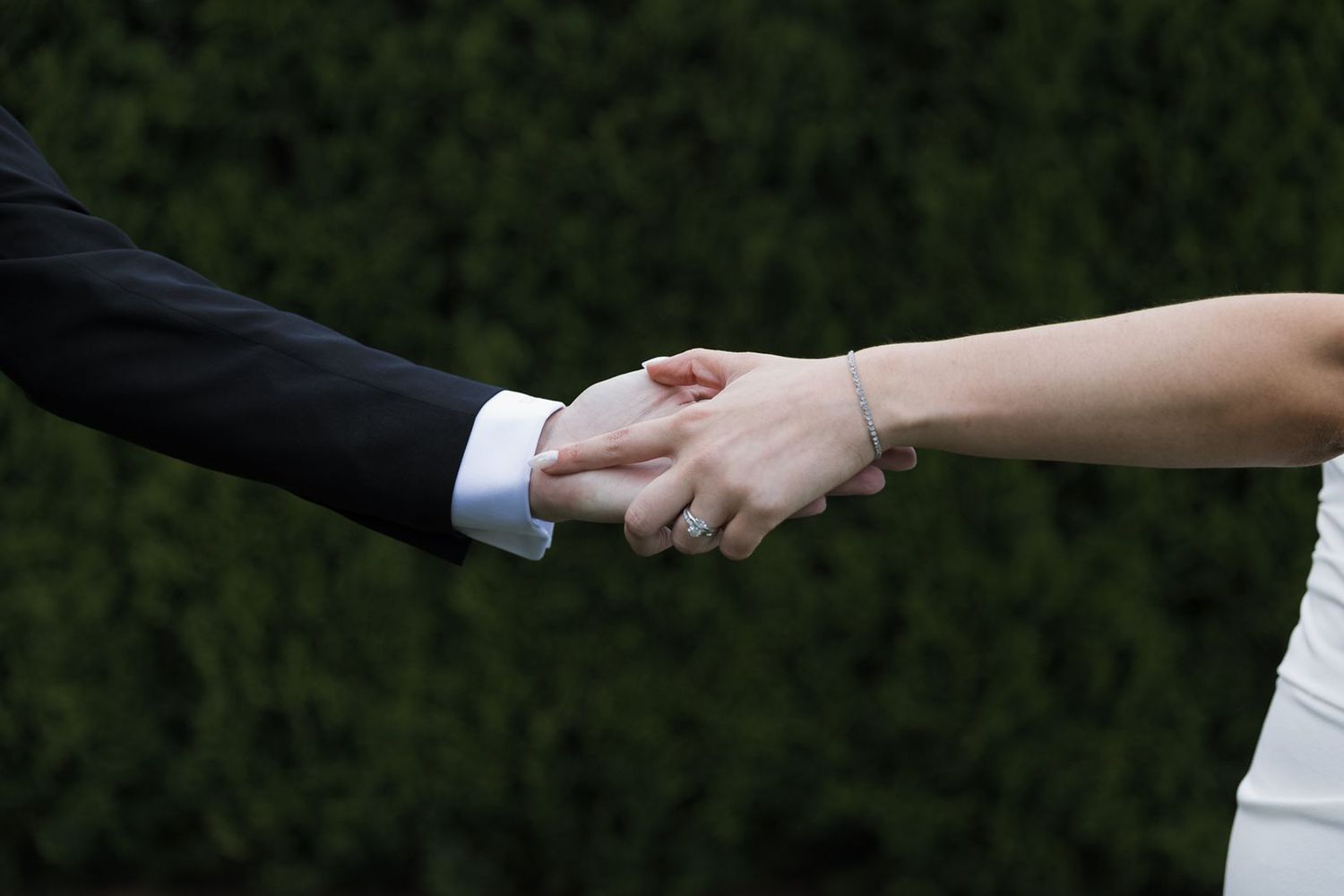 Close-up of bride and groom holding hands showing wedding ring and bracelet against green hedge backdrop.
