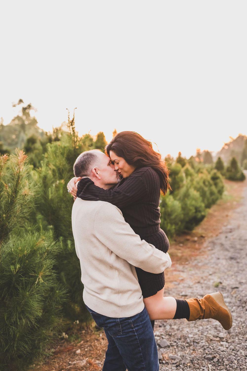 A romantic couple embraces and kisses on a rocky path surrounded by evergreen trees at sunset.