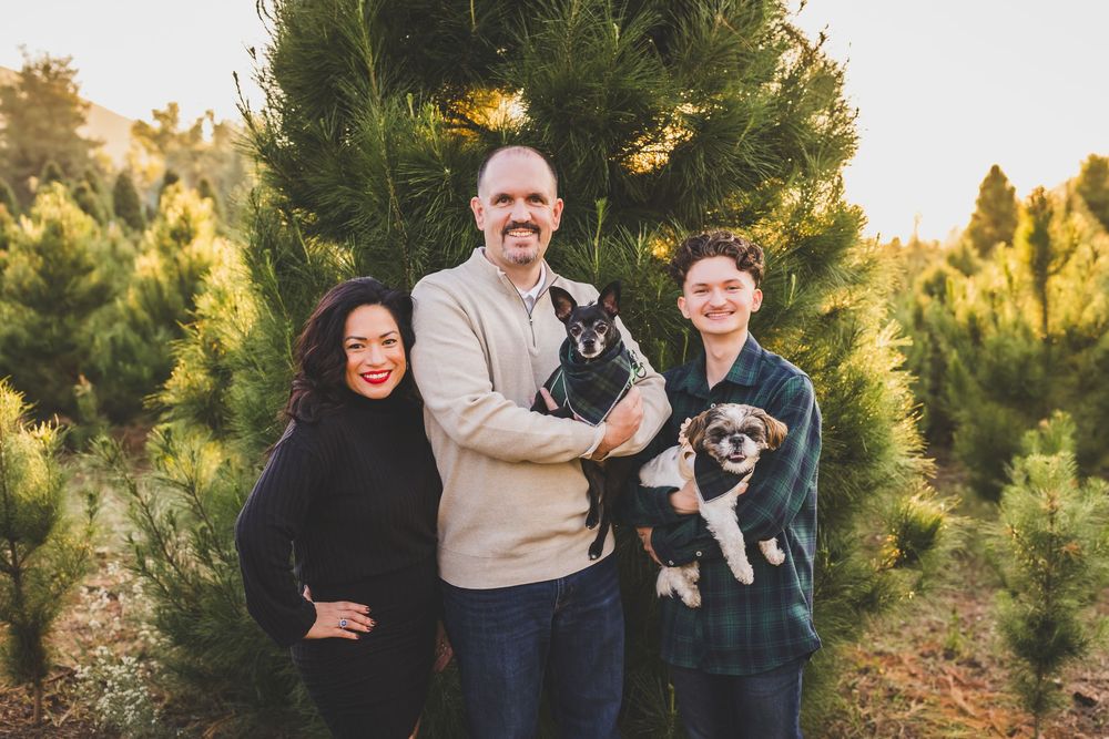 Family poses with their dog in a sunny outdoor setting with evergreen trees in the background.