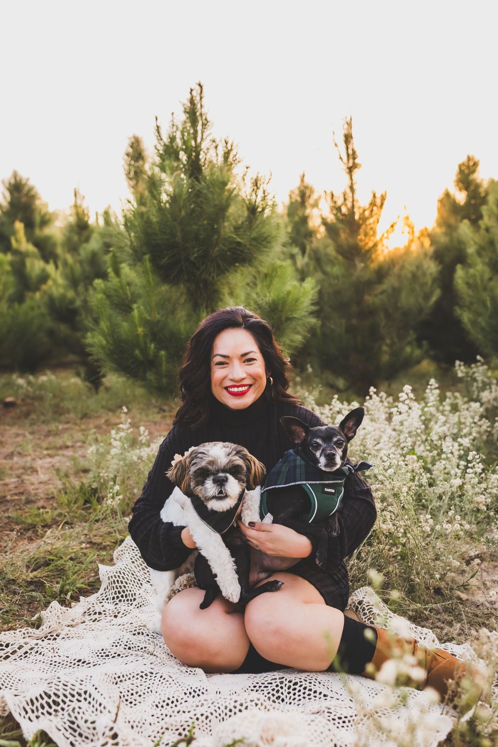 A person in black clothing sits on a white lace blanket outdoors with their small black and white dog.