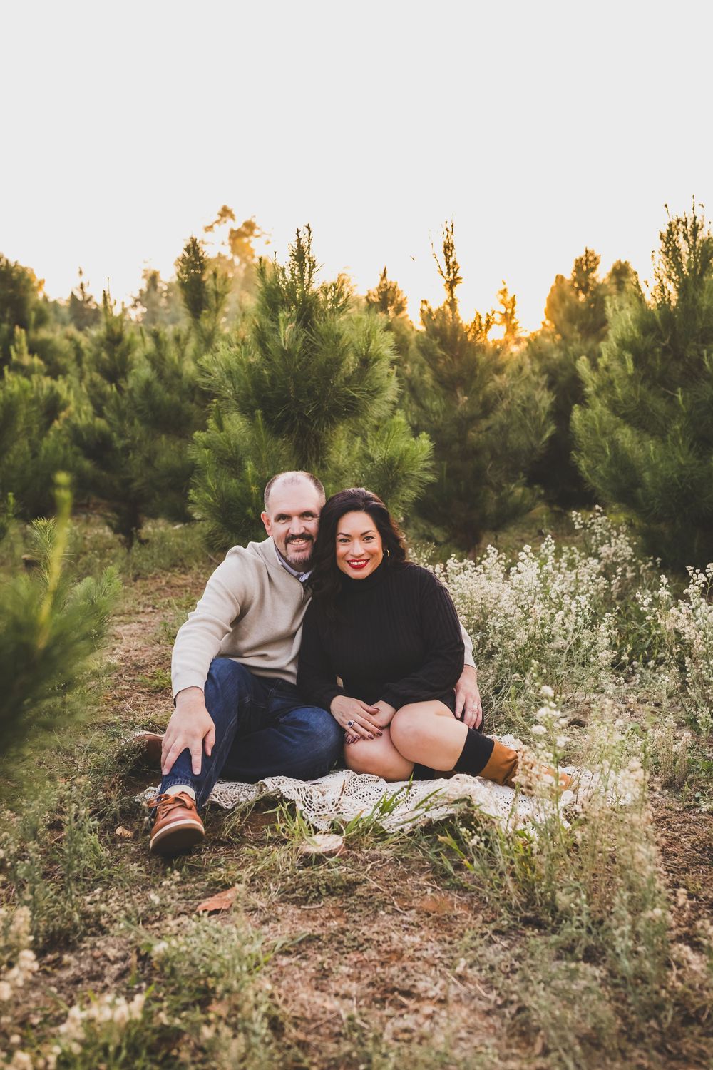 Couple relaxes on ground surrounded by pine trees and wildflowers during sunset photoshoot.