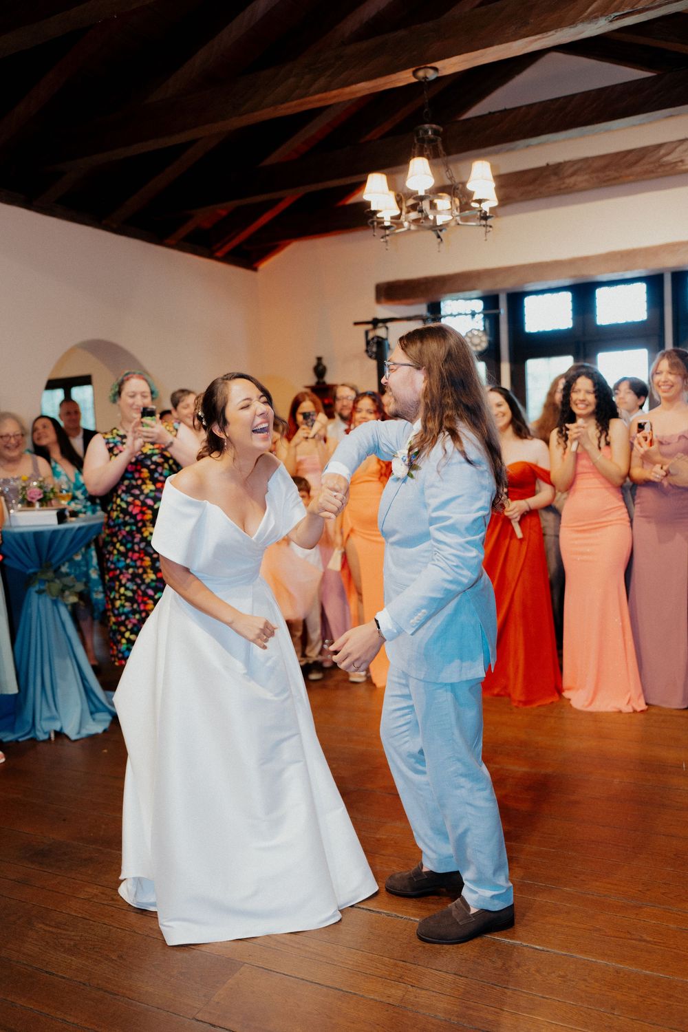 Wedding guests dance together on a wooden floor at a reception with colorful formal dresses and a chandelier overhead.