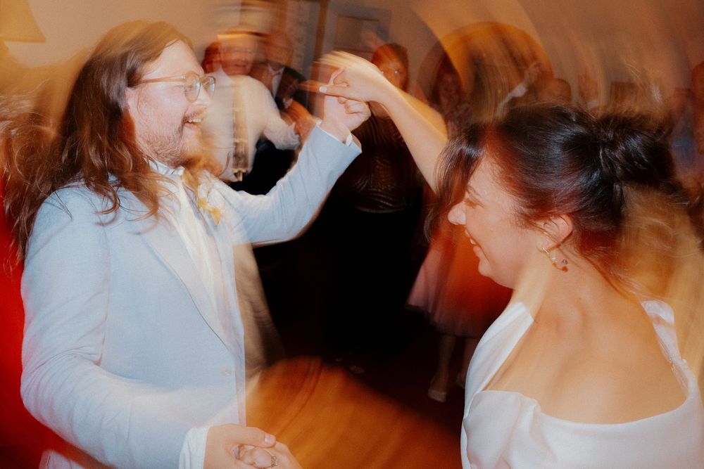 Wedding reception guests dancing and celebrating on a wooden dance floor under warm lighting.