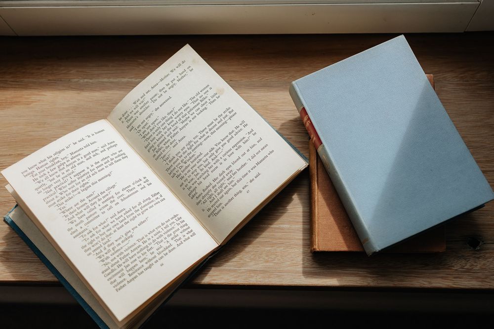 An open book with printed text lies on a wooden surface next to two closed hardcover books.