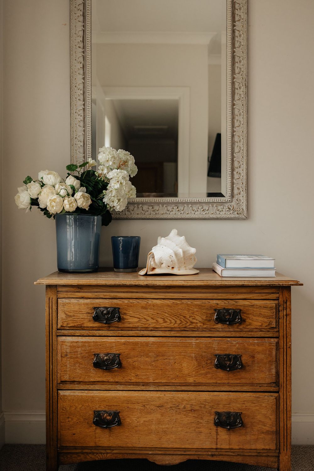 Vintage wooden dresser with ornate mirror displaying white hydrangeas in a blue vase and decorative accessories.