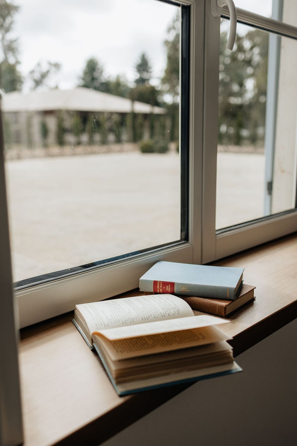 Open books resting on a windowsill with a blurred outdoor view through large glass windows.