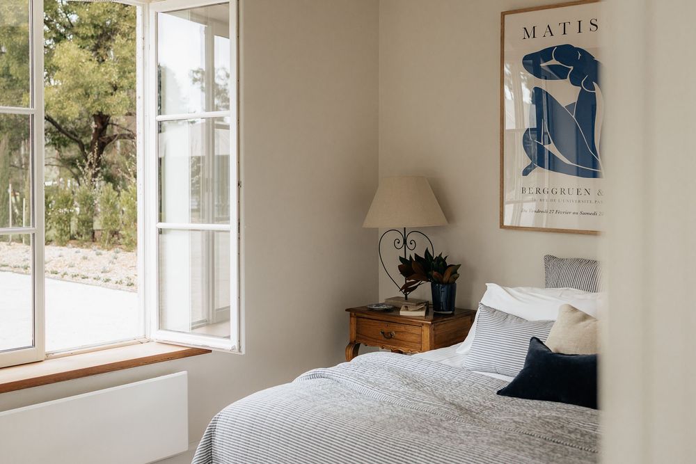 Minimalist bedroom interior featuring Matisse prints, wooden nightstands, and white linens with blue throw pillows.