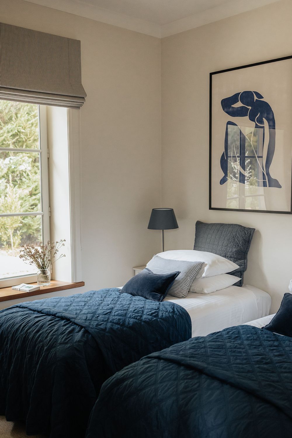 A minimalist bedroom with navy blue bedding and modern art prints features bright natural light from multiple windows.