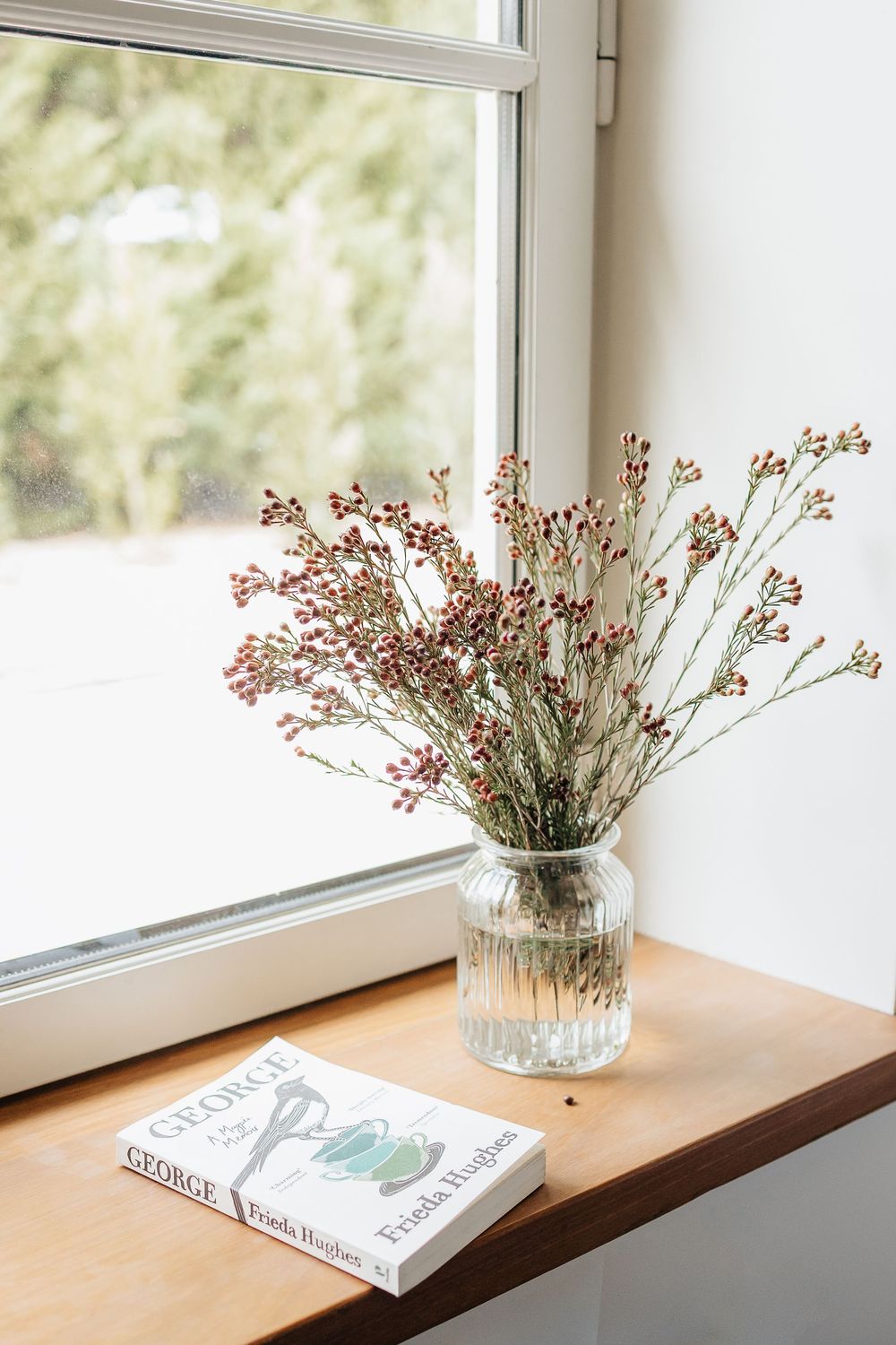 Dried pink flowers in a glass vase on wooden windowsill with book beside natural light.