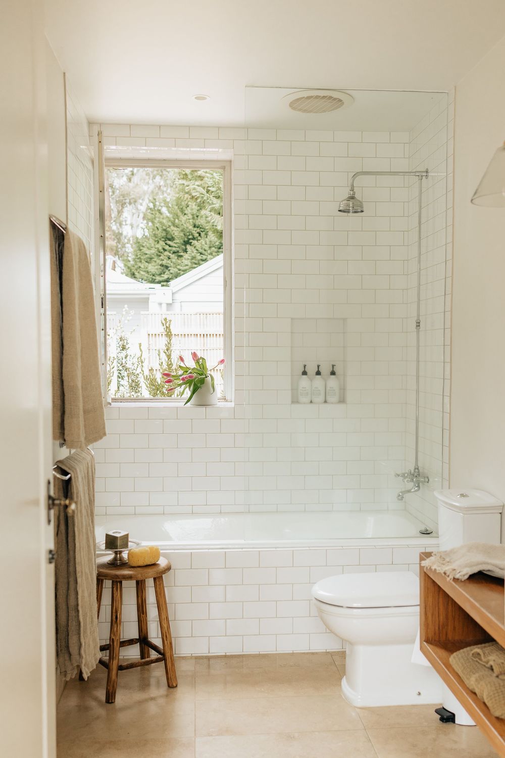 Modern white bathroom interior with subway tile walls, glass shower enclosure, wooden stool and natural light from window.