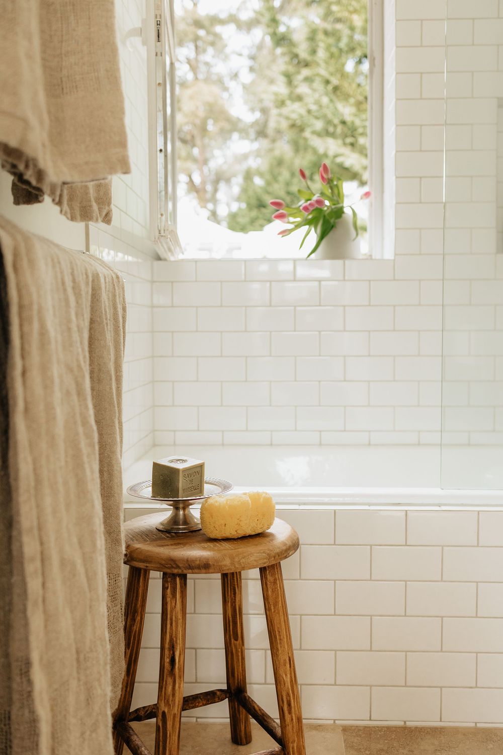 A wooden stool with a loofah and soap sits by a window with white subway tile walls and linen curtains.