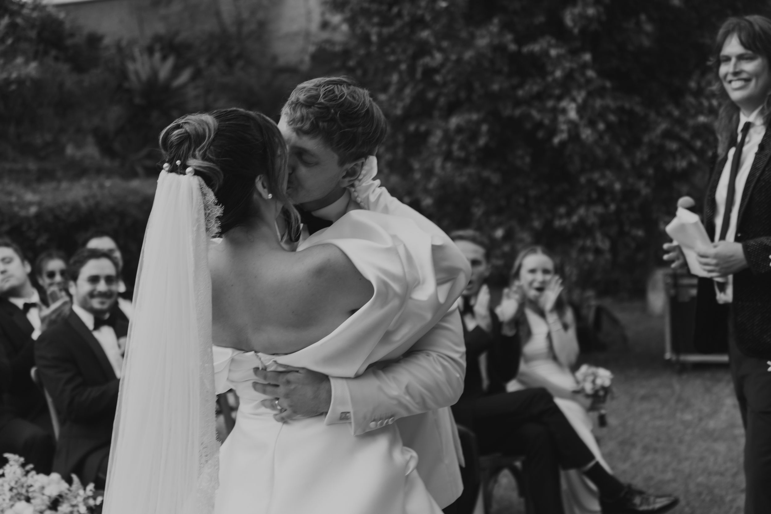 A black and white photograph captures a romantic first kiss between newlyweds at their outdoor wedding ceremony.