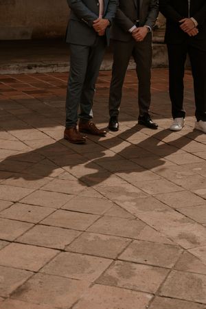 Three people wearing formal suits and dress shoes stand on stone tile flooring casting long shadows in warm lighting.