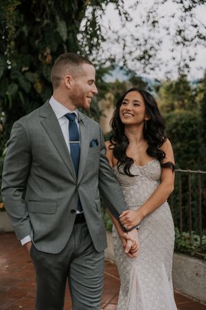 A romantic wedding couple shares joyful moments together in an outdoor garden setting wearing grey suit and lace gown.