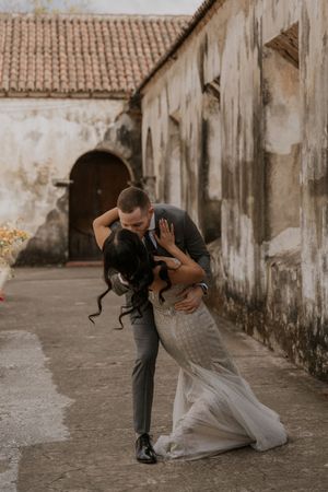 A couple shares an intimate dance moment in front of a weathered colonial building with arched doorways.