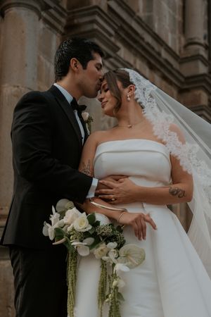 Romantic wedding couple embracing while holding white floral bouquet against stone architecture.