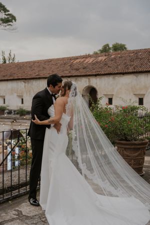 Newlyweds share an intimate moment on a terrace with rustic buildings in the background.