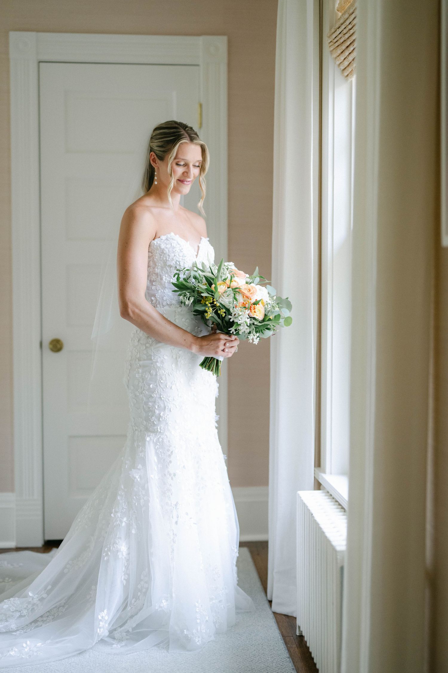 A bride in a white lace wedding gown holds a green and white bouquet while standing near sunlit windows. - Lake Forest