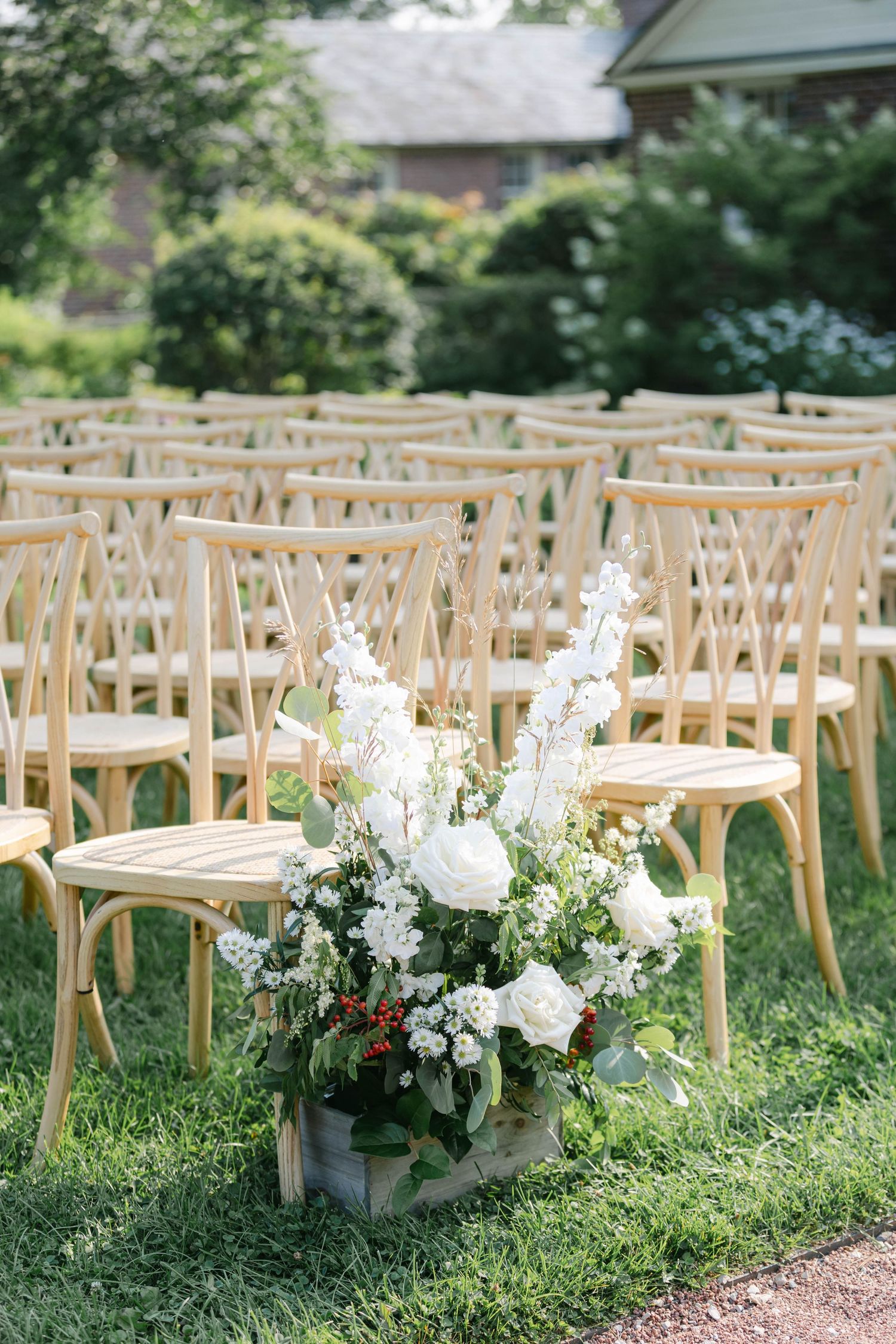 Rows of light wooden chairs arranged on grass with white floral arrangements lining the outdoor wedding ceremony aisle. - Elawa Farm