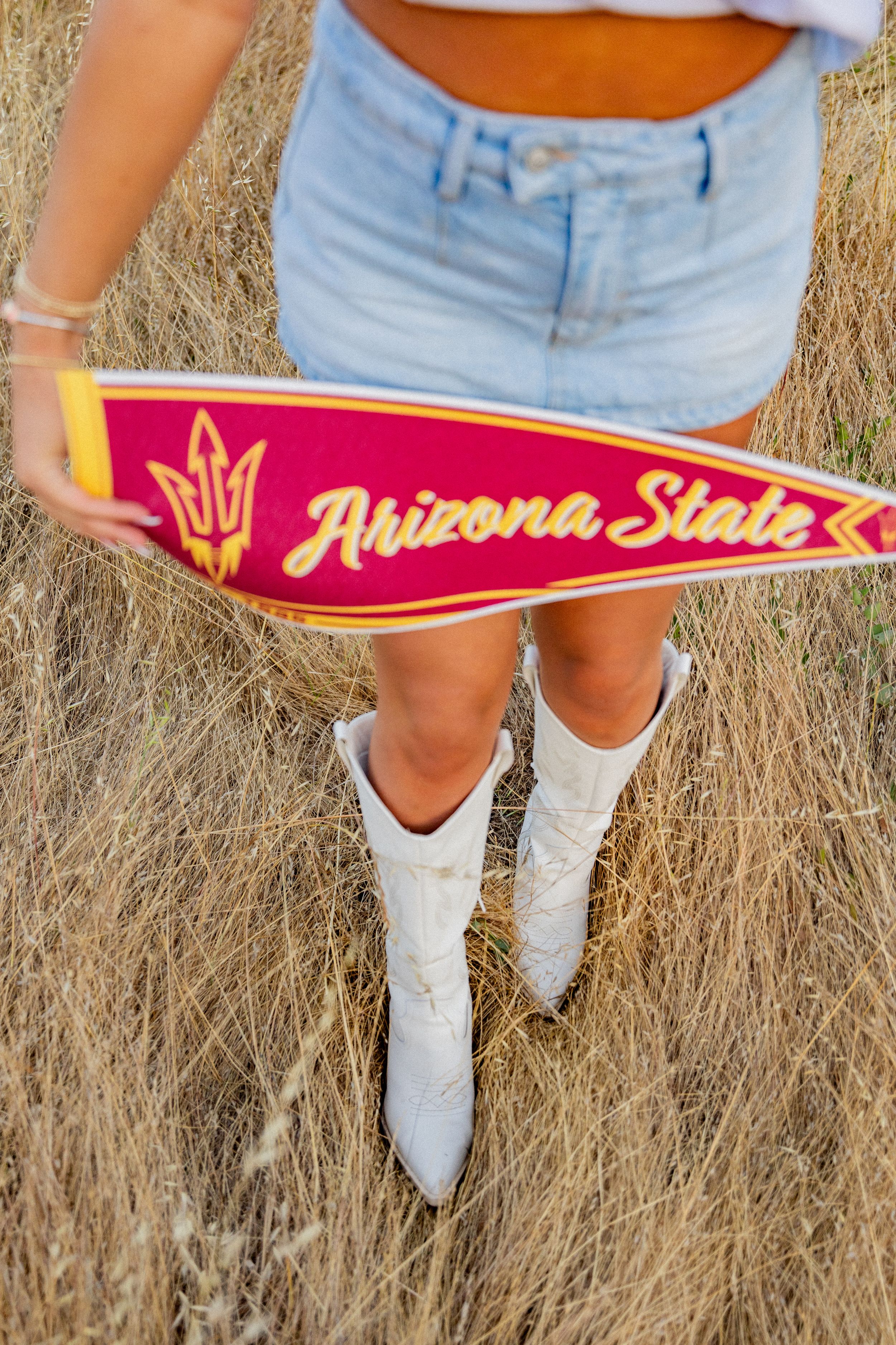 White cowboy boots and jean shorts against a dried grass field with an Arizona State University pennant.