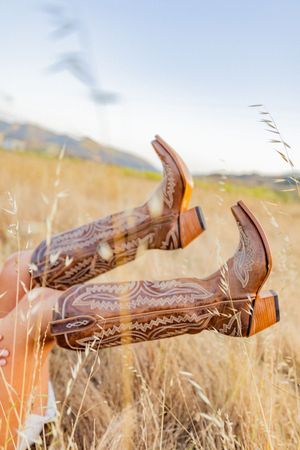 Decorative brown leather cowboy boots with intricate patterns shown against dry grass in a country field.