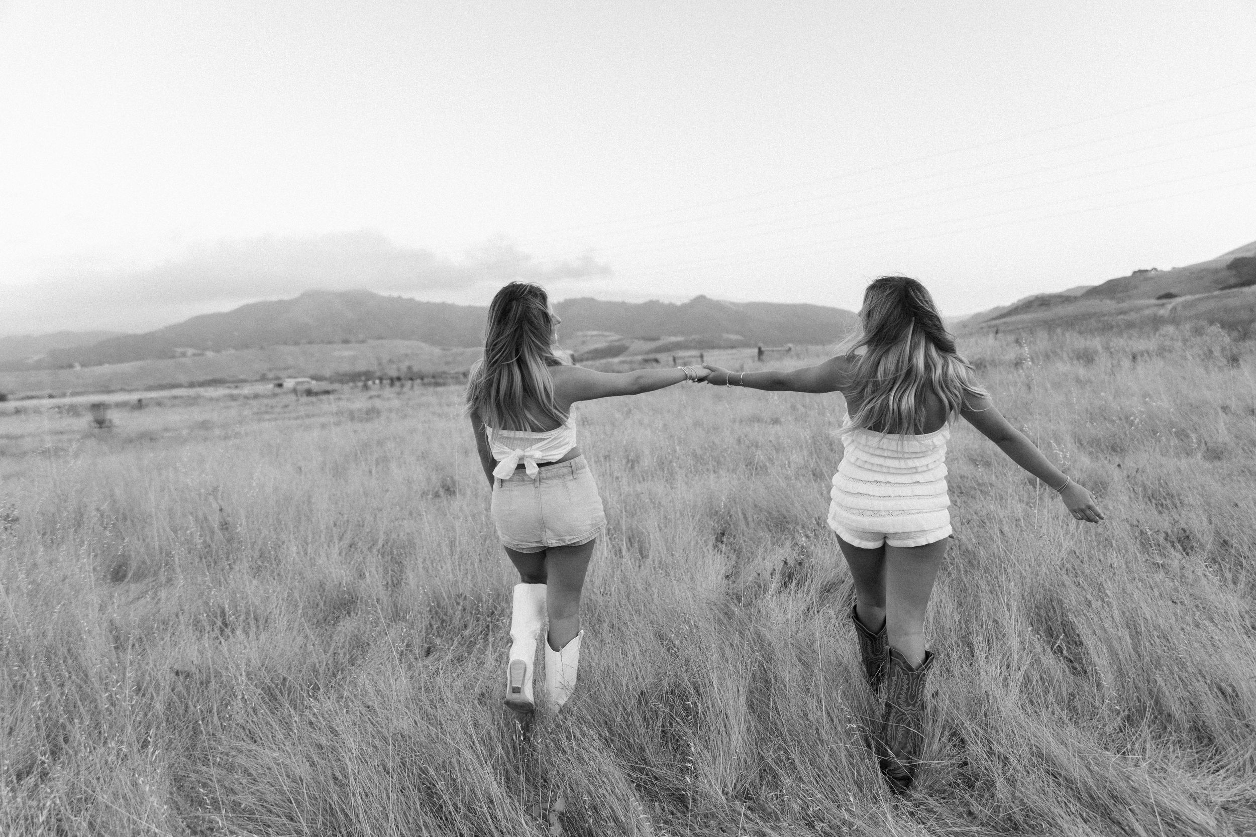 Two women walking through waist-high grass in an open field with mountains visible in the distance, shown in black and white.
