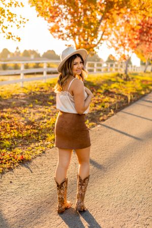 Full body fashion shot with cowboy boots and fall outfit against autumn trees and white fence.