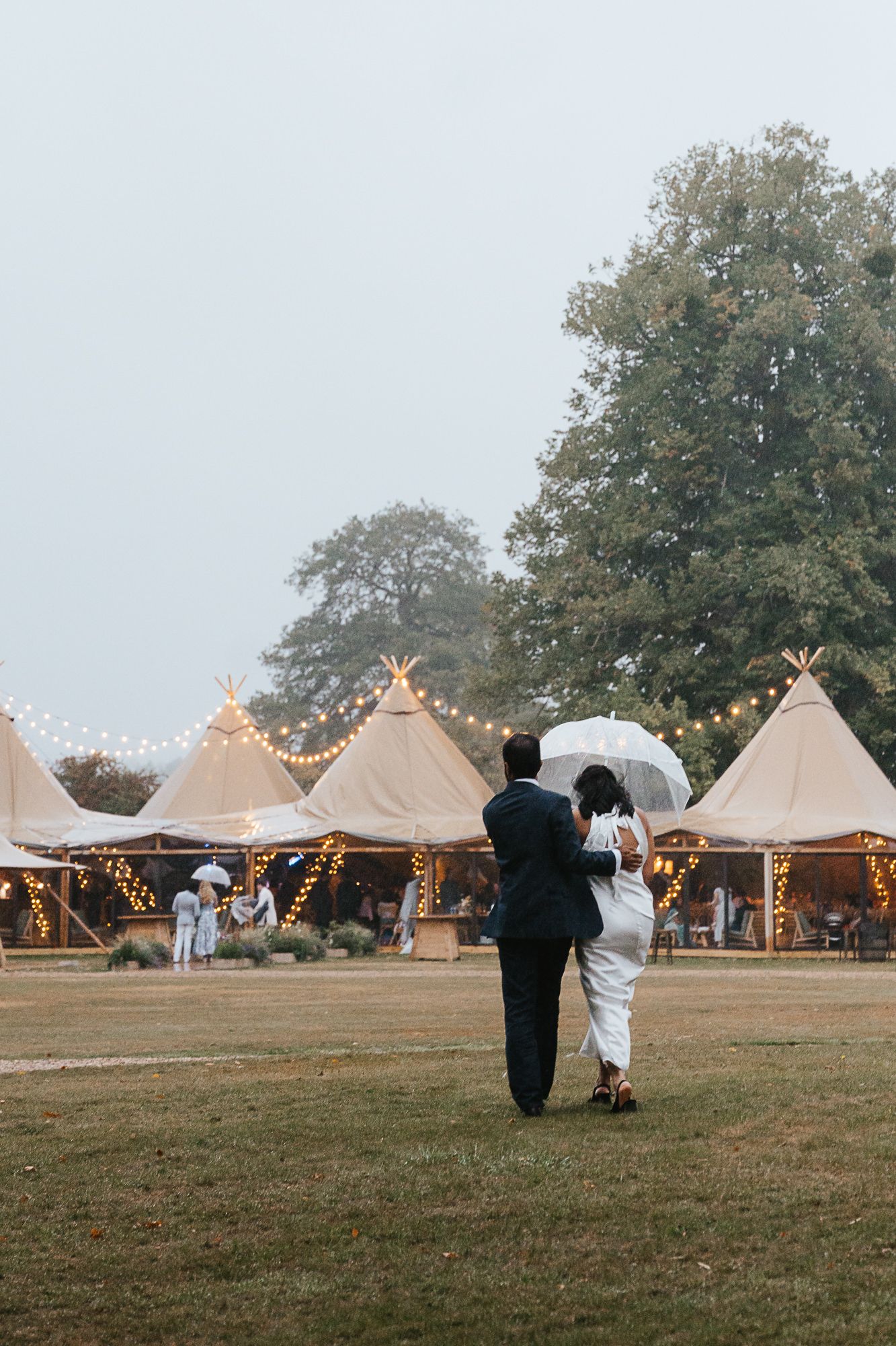 A couple walks toward illuminated tipi tents at a romantic outdoor wedding venue with string lights at dusk.
