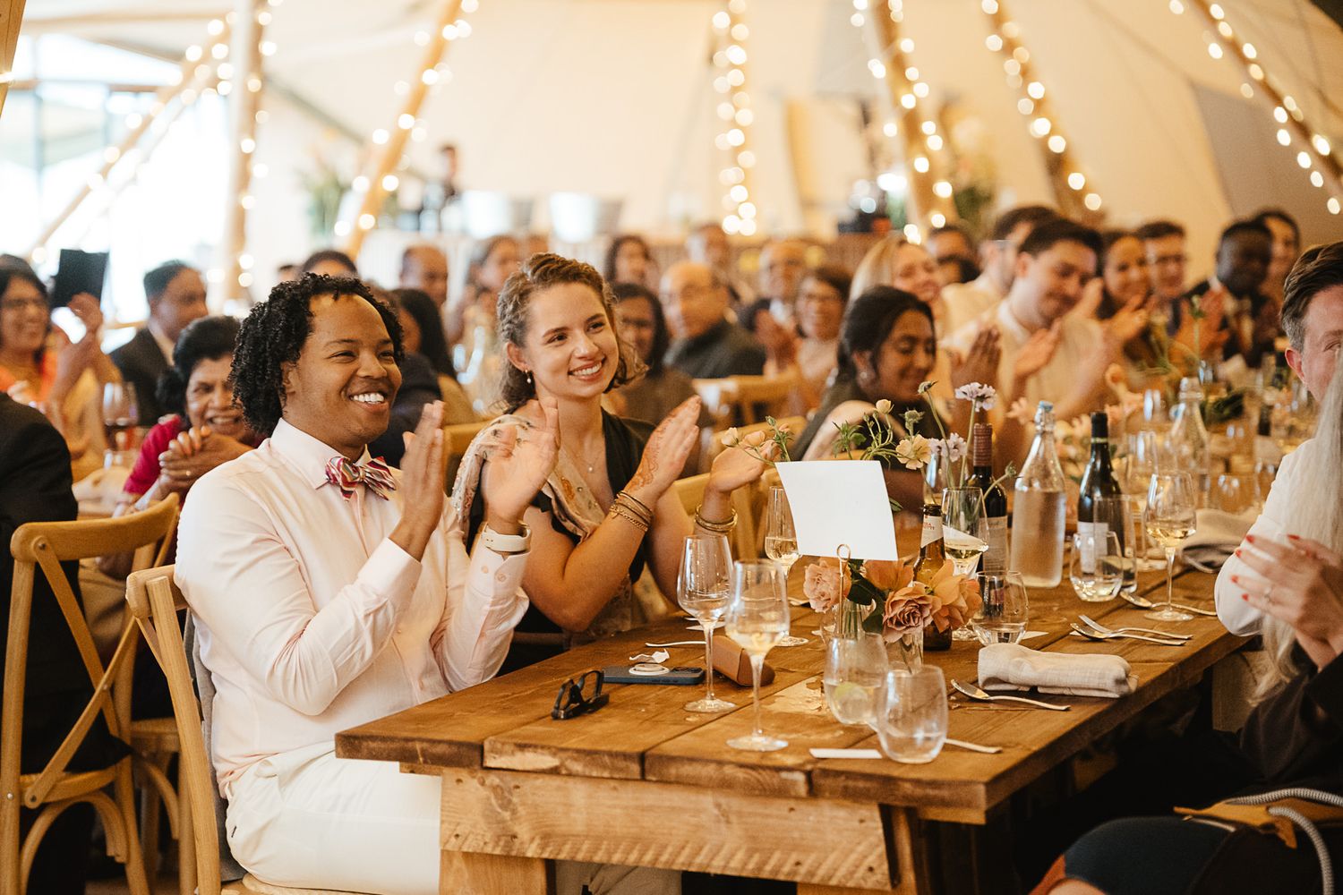 Wedding reception guests seated at long tables applaud during a celebration in a rustic venue with warm lighting.