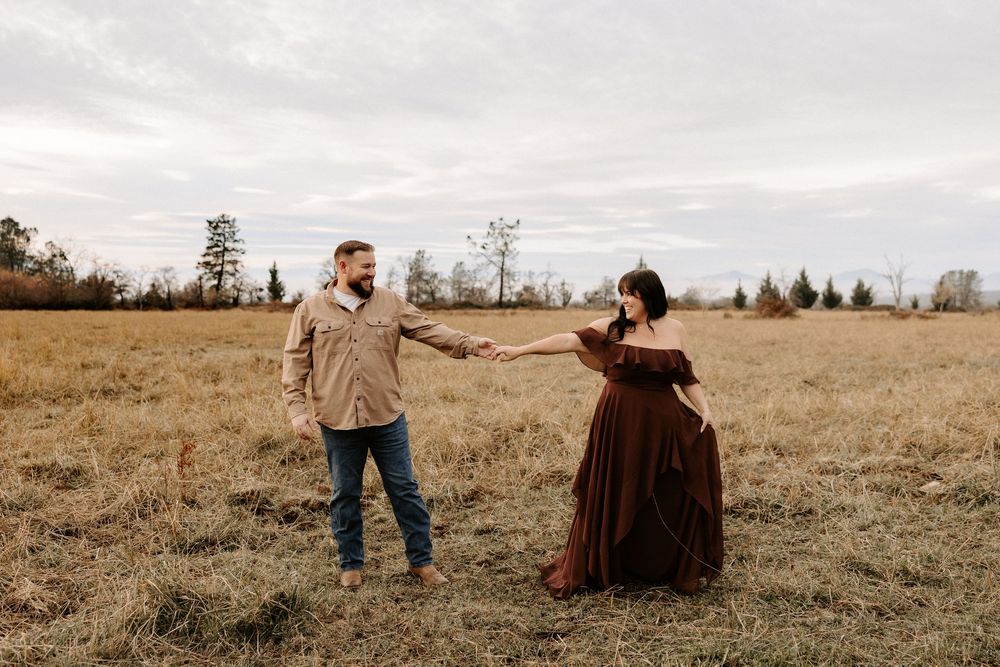 A couple sits together in a grassy field during golden hour as the sun sets in the distance.