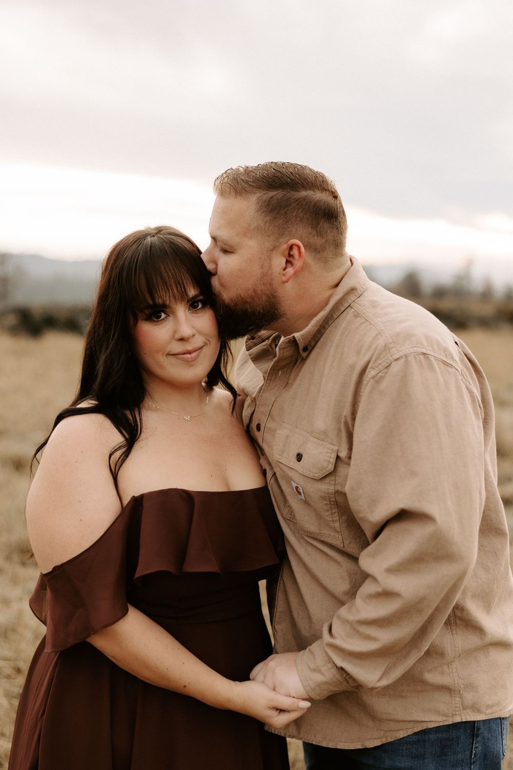Romantic couple embraces in a field during sunset, with her wearing a brown off-shoulder dress and him in a tan shirt.
