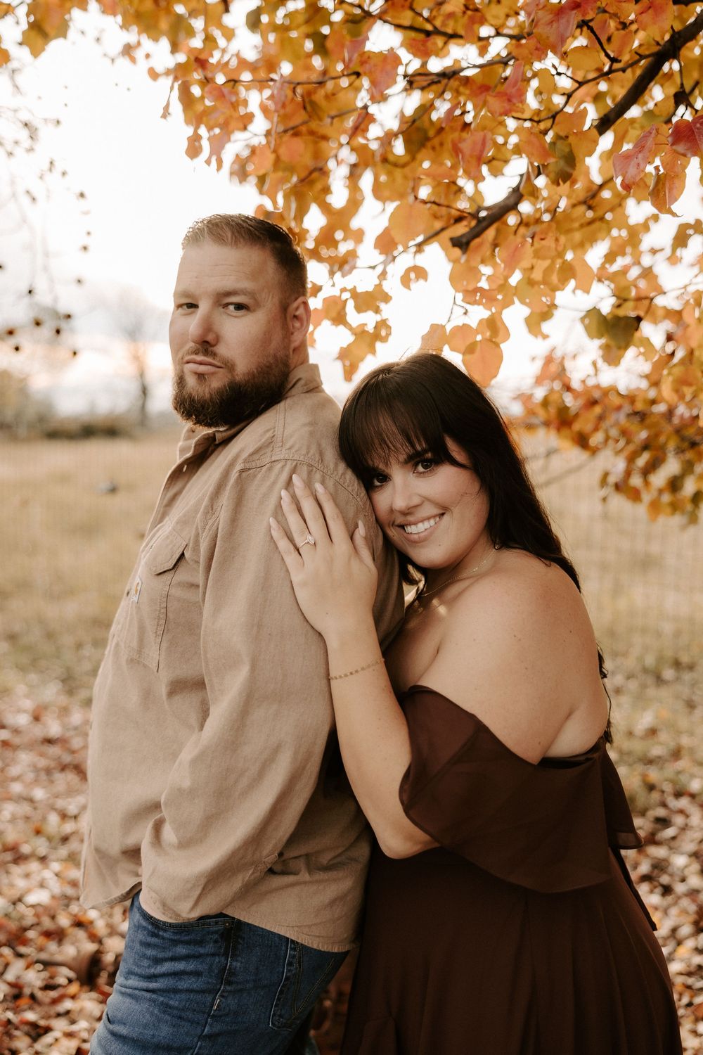 Couple poses beneath autumn leaves, with her in brown dress and him in tan shirt against a fall backdrop.